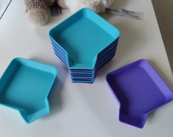 May include: A stack of turquoise and purple square-shaped trays with a unique cutout design. Two individual trays in turquoise and purple are also visible. The trays are made of a solid material and are stacked on a white surface.