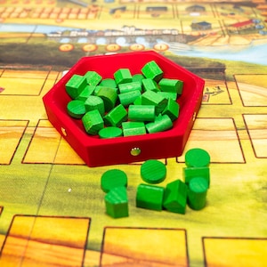 May include: A red, hexagonal tray filled with various green wooden game pieces, including cylinders, cubes, and rounded shapes. The tray sits on a colorful game board with a landscape design.