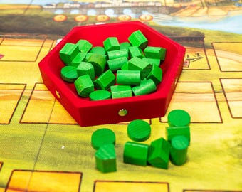 May include: A red, hexagonal tray filled with various green wooden game pieces, including cylinders, cubes, and rounded shapes. The tray sits on a colorful game board with a landscape design.