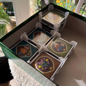 Puede incluir: Una caja de juego verde y marrón con divisores de tarjetas blancos. La caja contiene pilas de cartas de juego cuadradas con ilustraciones coloridas. Las cartas presentan imágenes de personajes y objetos.