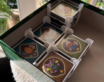 May include: A green and brown game box with white card dividers. The box contains stacks of square game cards with colorful illustrations. The cards feature images of characters and objects.
