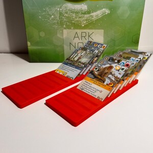 K&ouml;nnte beinhalten: Zwei leuchtend rote Kartenhalter mit mehreren Spielkarten. Die Karten haben verschiedene Illustrationen und Text. Der Hintergrund zeigt eine gr&uuml;ne Box mit dem Text "ARK NOVA".