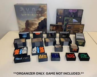 May include: A board game organizer set with multiple black storage boxes and containers. The set includes boxes for cards, tokens, and other game components. The game box for "Dune: Uprising" is visible in the background. The organizer is designed to keep game pieces organized.