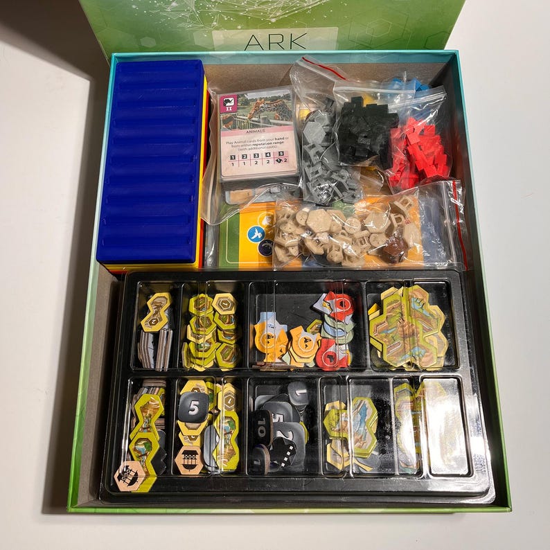K&ouml;nnte beinhalten: Ge&ouml;ffnete Brettspielbox mit verschiedenen Komponenten. Enth&auml;lt ein blaues rechteckiges Teil, Karten und Kunststoff-Spielsteine in Rot, Schwarz und Beige. Die Spielsteine sind in einem schwarzen Tablett mit mehreren F&auml;chern angeordnet. Die Box ist gr&uuml;n und tr&auml;gt das Wort "ARK".
