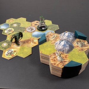Puede incluir: Un juego de mesa con fichas hexagonales que representan paisajes, montañas y un castillo. Una figura de monstruo verde y una miniatura de castillo están en el tablero de juego. Las piezas del juego están apiladas, listas para jugar.