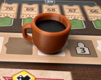 May include: A small, brown coffee cup filled with dark liquid, likely coffee, sits on a game board. The cup has a handle and is positioned near game pieces and numbered cards. The overall theme suggests a board game accessory.