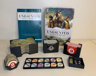 May include: A board game titled "Undaunted: Battle of Britain" with various game components. The game box and rule book are visible, along with black storage containers holding cards, tokens, and dice. The game components are arranged on a light-colored surface.