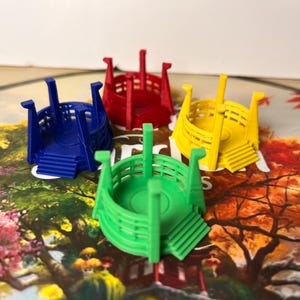 Puede incluir: Cuatro piezas de juego coloridas en azul, rojo, amarillo y verde, que se asemejan a pequeños botes o estructuras estilizadas. Cada pieza tiene una base circular con escalones y soportes verticales. Las piezas están dispuestas sobre un tablero de juego con un paisaje ilustrado colorido.