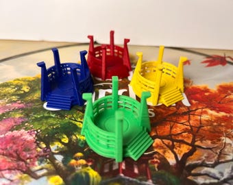 May include: Four colorful game pieces in blue, red, yellow, and green, resembling small, stylized boats or structures. Each piece has a circular base with steps and vertical supports. The pieces are arranged on a game board with a colorful, illustrated landscape.