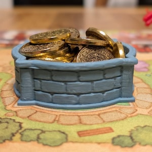 May include: A blue, brick-textured well filled with gold coins. The well is a small, decorative container, likely for a board game. The coins are shiny and golden, contrasting with the muted blue of the well.
