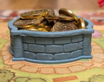 May include: A blue, brick-textured well filled with gold coins. The well is a small, decorative container, likely for a board game. The coins are shiny and golden, contrasting with the muted blue of the well.