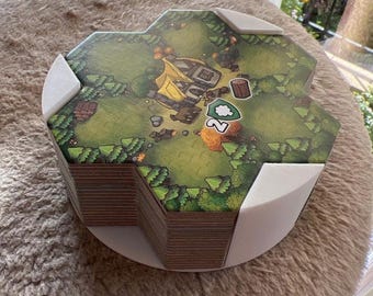 May include: A board game with hexagonal tiles featuring a forest scene with a campsite, trees, and a river. The tiles are stacked on a white circular base with white corner supports. The game appears to be a strategy or adventure game.
