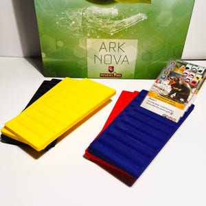 K&ouml;nnte beinhalten: Das Brettspiel "ARK NOVA" zeigt schwarze, gelbe, rote und blaue Spielkomponenten. Eine Spielkarte mit einer Illustration des Tasmanischen Teufels ist sichtbar. Die Spielkomponenten liegen auf einer wei&szlig;en Oberfl&auml;che.