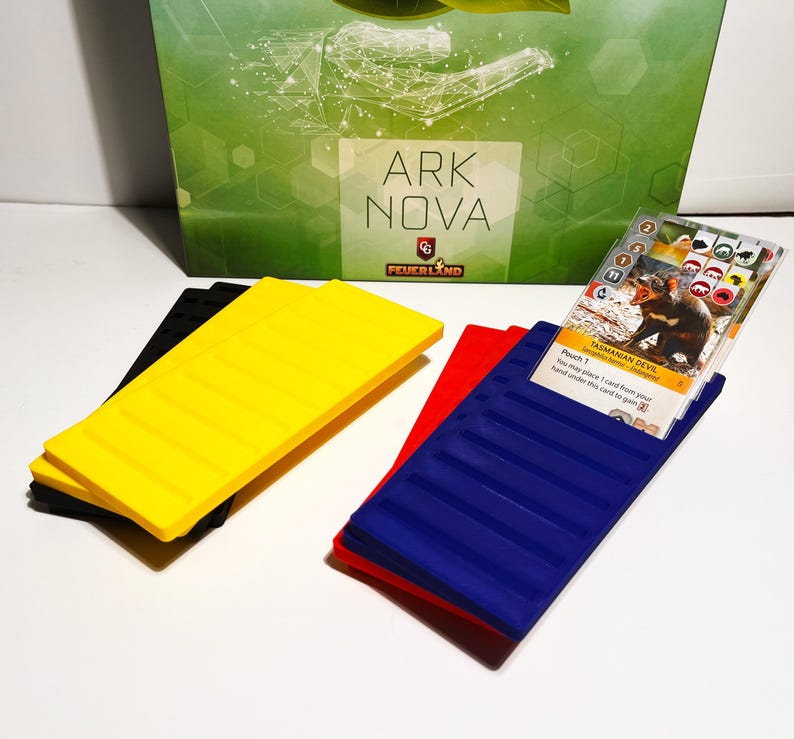 K&ouml;nnte beinhalten: Das Brettspiel "ARK NOVA" zeigt schwarze, gelbe, rote und blaue Spielkomponenten. Eine Spielkarte mit einer Illustration des Tasmanischen Teufels ist sichtbar. Die Spielkomponenten liegen auf einer wei&szlig;en Oberfl&auml;che.