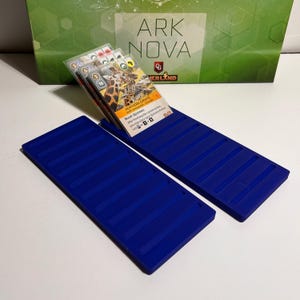 K&ouml;nnte beinhalten: Zwei blaue Kartenhalter aus Kunststoff mit gerilltem Design, die Spielkarten halten. Die Karten zeigen Bilder von Tieren und Text, darunter "Northern Giraffe". Im Hintergrund befindet sich eine gr&uuml;ne Box mit dem Text "Ark Nova".