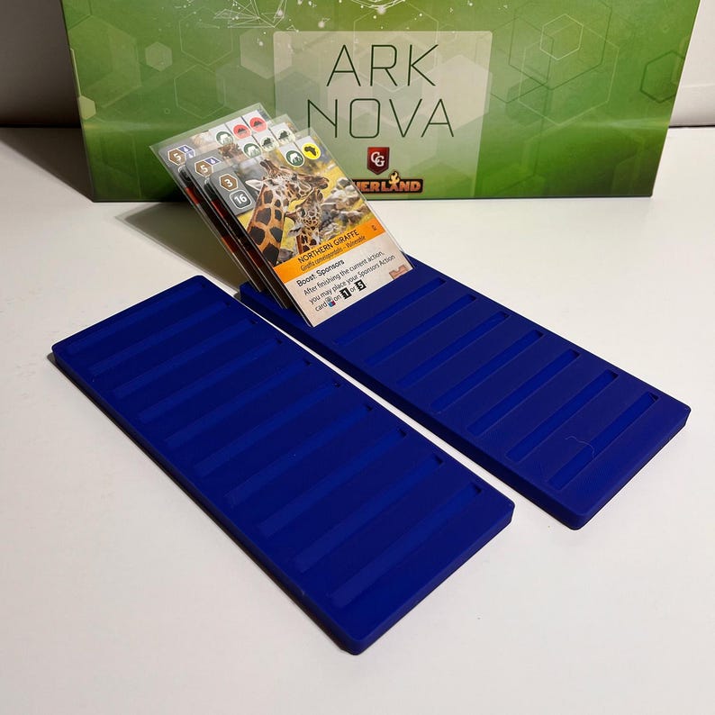 K&ouml;nnte beinhalten: Zwei blaue Kartenhalter aus Kunststoff mit gerilltem Design, die Spielkarten halten. Die Karten zeigen Bilder von Tieren und Text, darunter "Northern Giraffe". Im Hintergrund befindet sich eine gr&uuml;ne Box mit dem Text "Ark Nova".
