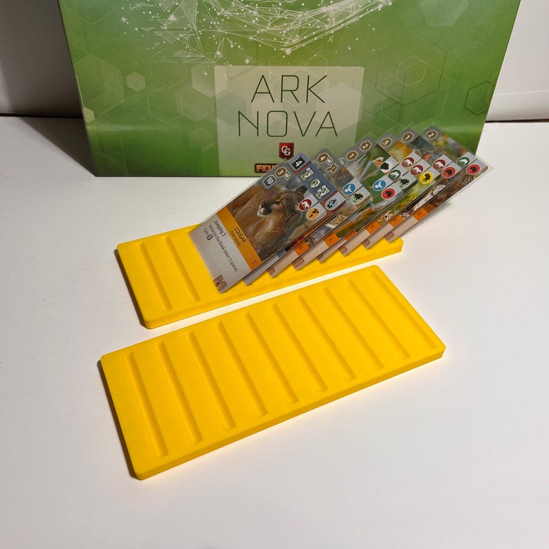 K&ouml;nnte beinhalten: Zwei leuchtend gelbe Kartenhalter mit einer Reihe paralleler Rillen. Ein Halter ist mit Spielkarten gef&uuml;llt. Die Karten zeigen Tierbilder und Symbole. Auf der Spielbox im Hintergrund steht der Text "ARK NOVA".