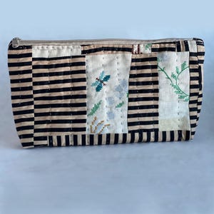 May include: A rectangular, zippered pouch with a patchwork design. The pouch features alternating black and tan stripes on the sides, and embroidered floral and insect motifs on the front. The zipper is a light beige color.