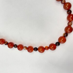 Peut inclure: Un collier composé de perles rondes de différentes tailles et textures, dans des tons de rouge, de marron et de noir. Le collier est présenté sur un fond blanc, mettant en valeur les couleurs des perles.