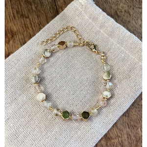 May include: A gold-toned bracelet with clear and green gemstone beads. The bracelet is on a beige linen pouch. The bracelet has a chain extension and a lobster clasp.