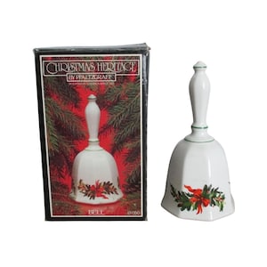 May include: A white ceramic bell with a handle and a green band, featuring a painted holly and red ribbon design. It is accompanied by a black rectangular box labeled Christmas Heritage by Pfaltzgraff with a picture of the bell on the front.