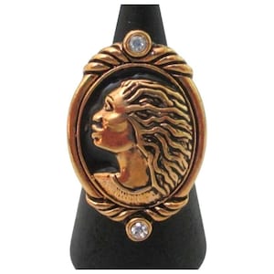 May include: An oval-shaped ring featuring a gold-toned metal cameo of a person in profile with wavy hair set against a black background. The frame is decorated with twisted metal detailing and two small, clear, round stones at the top and bottom.