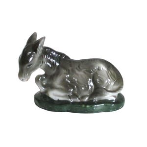 May include: A ceramic figurine of a donkey in a resting position. The donkey is gray with a white underbelly and is resting on a green base. The figurine has detailed features and a glossy finish, suitable for home decor.