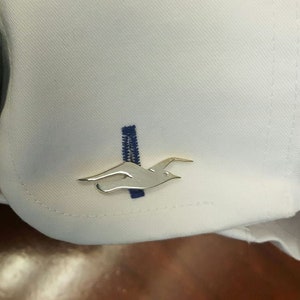 May include: A white shirt with a gold bird cufflink. The cufflink is a stylised bird in flight.