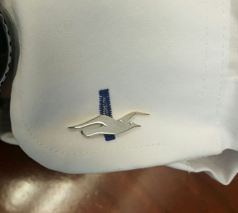 May include: A white shirt with a gold bird cufflink. The cufflink is a stylised bird in flight.