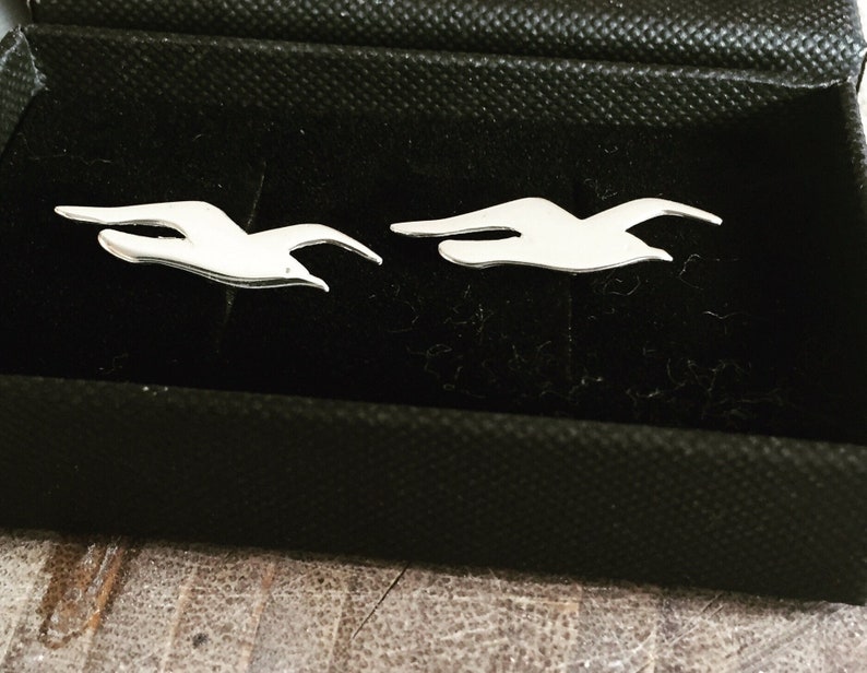 May include: Two silver bird cufflinks in a black velvet box. The birds are in flight and are facing the same direction.