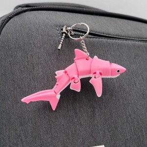 Puede incluir: Un llavero de tiburón articulado rosa y blanco colgando de un llavero plateado. El tiburón está sujeto a una mochila gris oscuro con un bolsillo con cremallera.
