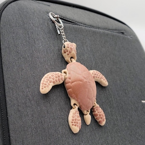 May include: A tan and brown turtle keychain hangs from a gray backpack. The turtle has a detailed design with articulated limbs and a textured shell. The keychain is attached with a silver chain.