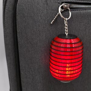 May include: A red, cylindrical lantern keychain with black accents hangs from a silver zipper pull on a dark gray backpack. The lantern is lit, and the backpack has a small pocket.