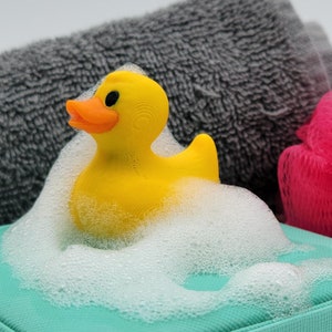 May include: A yellow rubber duck in a turquoise box with white foam, a grey towel, and a pink bath sponge.