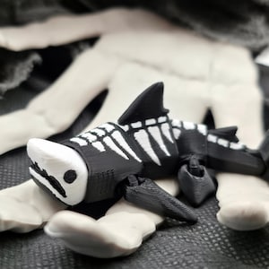 May include: A black and white shark skeleton figurine with a detailed rib cage and fins. The shark is resting on a light-colored, textured surface, with a dark gray background. The figurine has a unique, artistic design.