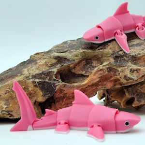 May include: Two articulated pink toy sharks are displayed on a piece of brown, textured rock. The sharks have a segmented body, fins, and a tail. One shark is positioned on top of the rock, while the other is at the base.