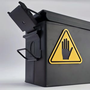 Könnte beinhalten: Eine schwarze Metallbox mit einem Klappdeckel und einem gelben Warnschild mit einem schwarzen Handsymbol. Die Box hat einen Griff an der Seite und ist so konzipiert, dass sie sich von oben öffnen lässt. Die Box ist rechteckig.