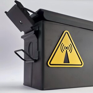 Könnte beinhalten: Eine schwarze Munitionskiste aus Metall mit einem aufklappbaren Deckel, der schräg geöffnet ist. Ein gelbes Dreieck mit einem Funksymbol ist auf der Vorderseite angebracht. Die Kiste hat einen Griff und einen Verschluss.