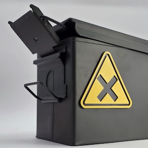 Könnte beinhalten: Schwarze Munitionskiste aus Metall mit aufklappbarem Deckel. Auf der Vorderseite befindet sich ein gelbes Gefahrensymbol mit einem schwarzen X. Die Kiste hat einen Griff und steht vor einem neutralen Hintergrund.
