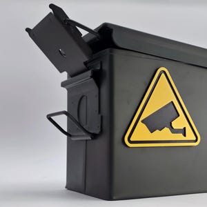 May include: A dark grey metal storage box with a hinged lid, displaying a yellow warning sign with a security camera symbol. The box has a handle and the lid is slightly ajar.