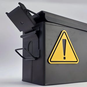 May include: A black metal ammunition box with a yellow warning sign. The box has a hinged lid that is partially open, revealing the interior. The warning sign is a yellow triangle with a black exclamation point.