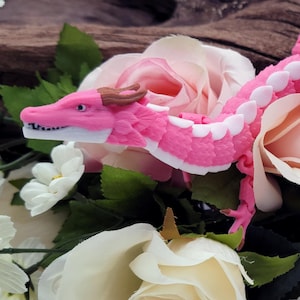 Puede incluir: Una figurita de dragón articulada de color rosa con detalles blancos, posada sobre un lecho de flores blancas y rosas.