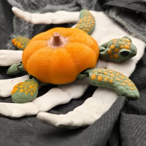 May include: A Halloween-themed decorative piece featuring a bright orange pumpkin surrounded by green and yellow-spotted appendages, resting on a white, skeletal hand. The background includes dark gray fabric and tattered cloth, creating a spooky atmosphere.