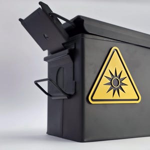 May include: A black metal storage box with a hinged lid, displaying a yellow warning triangle with a sunburst symbol. The box has a handle and the lid is slightly ajar. Likely for storing items.