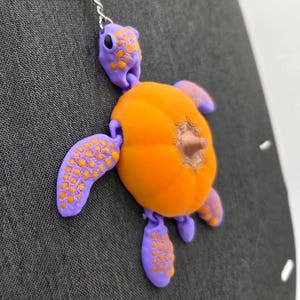 May include: A whimsical turtle charm in orange and purple, hanging against a gray fabric background. The turtle has a textured shell and flippers, with a small brown detail on its body. A silver chain holds the charm.