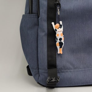 May include: A blue backpack with a black strap and a small, white, black and orange cat-shaped keychain attached.