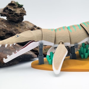 May include: A tan and white articulated Mosasaurus model with green accents, displayed on a yellow base with green coral and a fossil. The model has a detailed head and open mouth, showcasing its teeth. The background includes rocks.