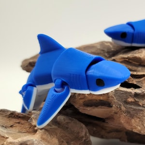 May include: Two blue articulated shark figurines with white accents, posed on a brown rock surface.