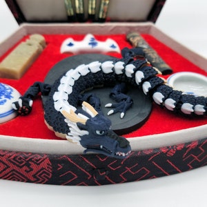 Puede incluir: Una figurita de dragón negro y blanco con detalles dorados se encuentra en una caja roja y negra con otros suministros de caligrafía. El dragón está enroscado alrededor de una piedra de tinta negra.