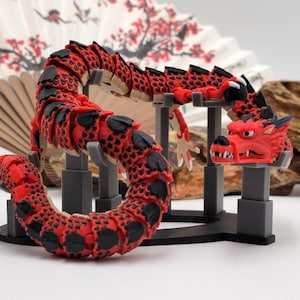Puede incluir: Una figurita de dragón articulada roja y negra en un soporte. El dragón está en posición enroscada con la cabeza levantada.